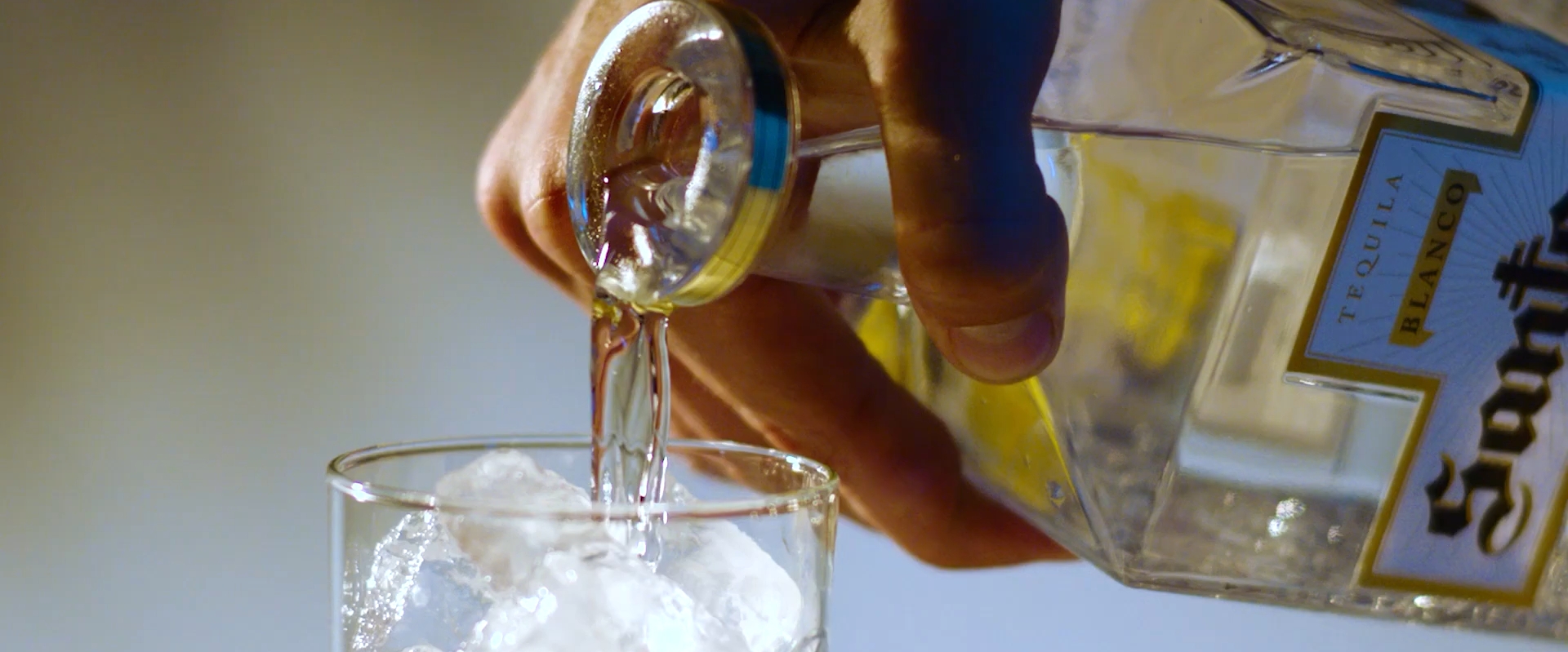 Hand holding a bottle of Santo Blanco tequila and pouring some into a glass of ice.